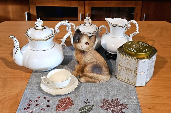 A tea tableau for National Cat Day with a large stonecast cat at the center, gazing limpidly into the camera. It is surrounded by an elegant, antique white teapot, creamer, and lidded sugar bowl with gold bead edging, a vintage white cup & saucer, and a white & gold tea tin of Harney and Sons Gingerbread Festival black tea. It all rests upon a grey linen placement embroidered with autumn leaves in muted earth tones.
