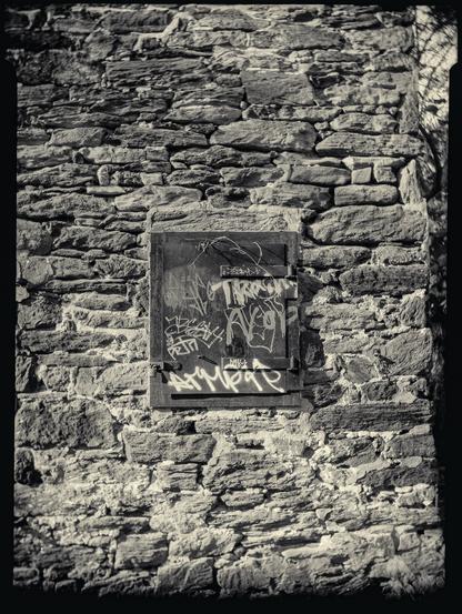 Black and white photo of a rusty hatch covered in graffiti in an old stone wall.