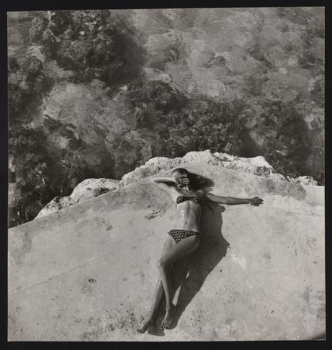 The image is a black and white photograph featuring a woman wearing a polka dot bikini. She appears to be lying down on the edge of an elevated surface, with her arms outstretched towards what seems like water below. The background consists of rough textures that could represent rocks or coral near the shore. There's no other person visible in the image and it evokes a sense of solitude or relaxation. This photograph is identified as Fashion model Natahli (Natalie) Nickerson Paine wearing a bikini, lying on platform near water, Montego Bay, Jamaica [1946 November].