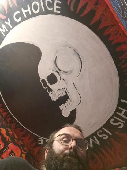 a selfie of a bearded man with glasses looking to the camera, on the ceiling the logo of the band Dead Moon.
it's a white skull in the shape of a cresent moon in white, while the beyond is black. in white Letters is written around: This is my choice, this is my voice