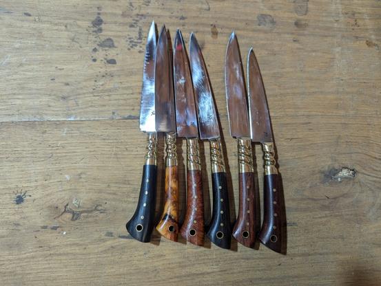 Six couteaux de table, de forme plutôt Renaissance, tous ont la même forme, avec des mitres torsadés et des rivets en laiton, chaque manche est fait d'une essence de bois différente, ils sont posés sur l'établi.