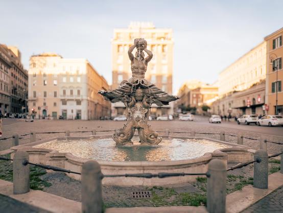 La Fontana del Tritone, una famosa fontana situata a Roma, Italia. Presenta una scultura di Tritone, un dio del mare della mitologia greca, raffigurato mentre tiene una conchiglia. La fontana si trova in un contesto urbano con edifici sullo sfondo.