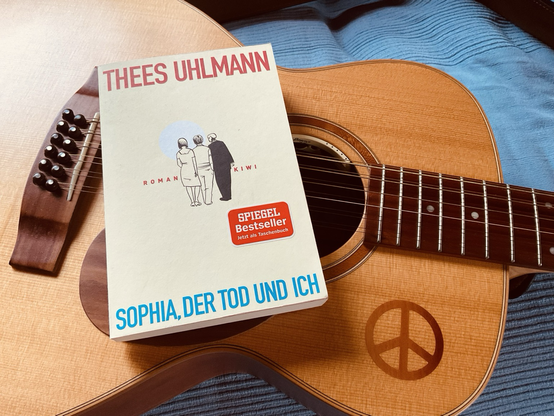 das buch „sophia, der tod und ich“ von thees uhlmann liegt auf einer 12saitigen gitarre der fa. lakewood mit in die decke eingearbeitetem peace-zeichen