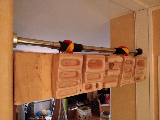 A fingerboard attached to a plywood plank, with two red hooks hanging from a pull-up bar in a doorway. The hooks are held in place by two yellow straps.