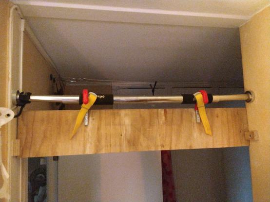 The back of the plywood plank. The yellow straps holding the red hooks on place on the pull-up bar is more visible. On each side of the doorway is a small piece of plywood attached to the wall. They prevent the whole contraption from swinging.