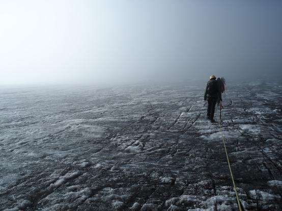 Sur un glacier gris et strié, des personnes avancent casquées dans le brouillard. On voit la corde de sécurité qui arrive jusqu'à moi qui suis hors champ.