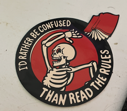 Circular sticker showing a skeleton throwing a red hardback book. Text around the image says, “I’d rather be confused than read the rules”