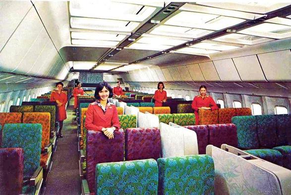 6 smiling flight attendents stand and pose for the camera. They are inside the plane with some very colorful seats.