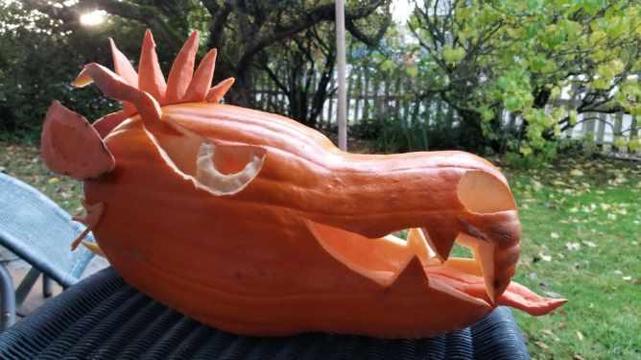 Side profile view of dragon jack-o-lantern.
Sitting on black wicker table. Background is backyard with fruit trees & fallen Autumn leaves.