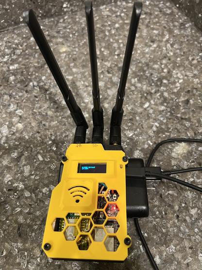 A bright yellow rectangular wardriver with three large black antennas has flashing lights and a blue display.