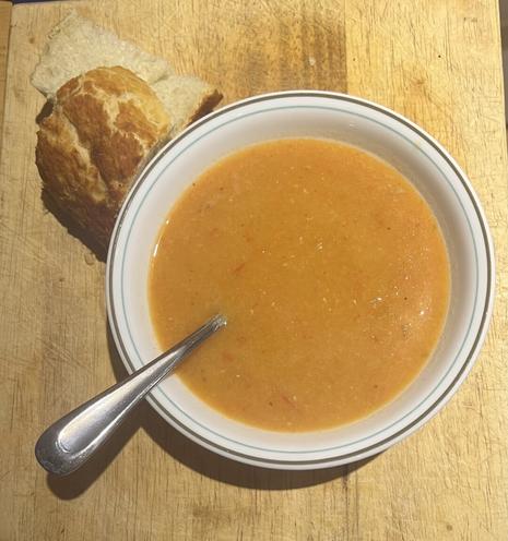 bowl of red lentil soup on a wooden surface, soup bowl has a spoon in it and to the top left there is a cut piece of crusty bread just next to the bowl.