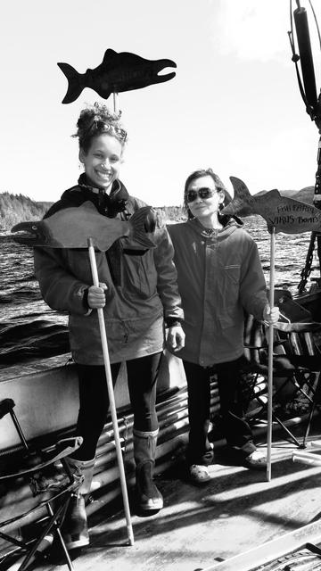 Me & a black female activist on a large Indigenous owned fishing boat, holding our fish farms protest signs. The signs are salmon themed & mounted on wooden sticks.
Black and white photo.