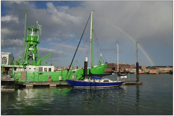 Ancien bateau phare reconverti en PUB dans le port de Gosport, UK. Le bateau, encore surmonté de son phare, est peint en vert. Amarré sur un ponton flottant qu'il partage avec un voilier bleu. Au premier plan, l'eau sombre reflete les couleurs des bateaux et du ciel. Celui-ci est chargé de nuages sur lesquels se dessine un arc-en-ciel qui témoigne de la météo incertaine.