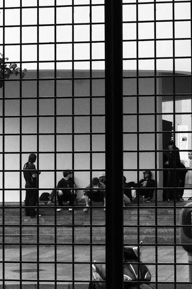 A group of persons sitting on skateboards or standing on stairs below an overhanging structure that shelters them from the rain. Photographed through a steel grating door.