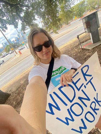 Selfie of a woman in sunglasses and a "Rethink I-35" shirt standing in a sprawlscape with tall buildings in the background, holding a hand-written sign reading "Wider Won't Work"