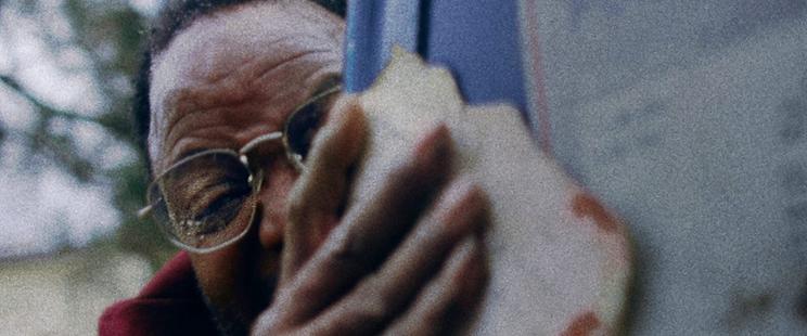 Still from the film DEAD MAIL. A man's close-up face in distress. He is half obscured by a mailbox in the foreground, and he grasps a bloody and torn piece of paper in his hand.