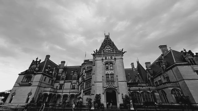 Black and white view of front of large Châteauesque-style mansion with cloudy sky above. The appearance is similar to scary black and white movies seen around Halloween.