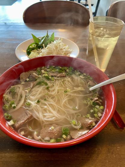 Steaming pho noodle soup in a large red bowl. Behind the bowl there is a plate of accompaniments, bean sprouts, lime wedge and Thai basil leaves. A glass of iced water with a straw can be seen beside the whole meal