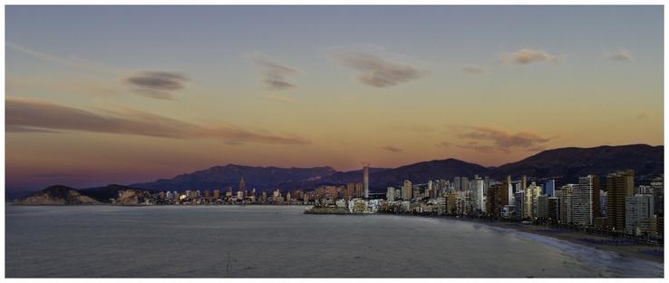 A panoramic view of a coastal city at sunrise. The image features a skyline of tall buildings along a beach, with mountains in the background and soft clouds in the sky. The colors of the sky transition from golden to purple as night turns to day.