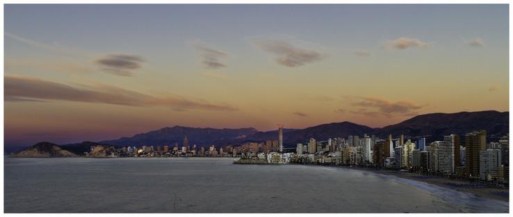 A panoramic view of a coastal city at sunrise. The image features a skyline of tall buildings along a beach, with mountains in the background and soft clouds in the sky. The colors of the sky transition from golden to purple as night turns to day.