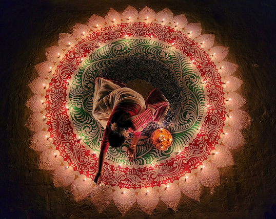 Looking directly down at an Indian woman in a decorative circle painted onto the ground. There are candles placed throughout the painted pattern that she is lighting. They shine brightly in the dark space around her.