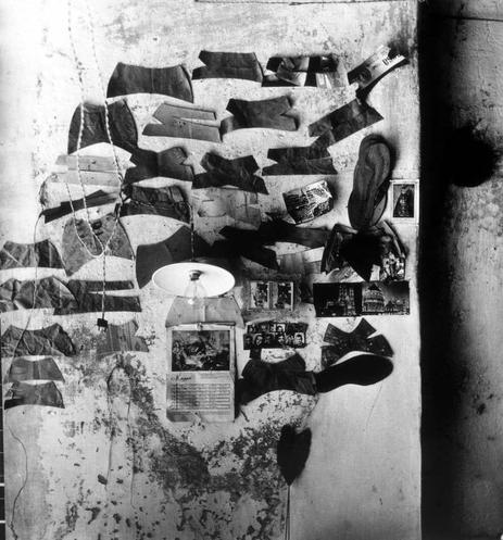 The image is a black and white photograph that appears to be taken from an overhead perspective, showcasing various items scattered on what seems like a floor or table. These objects include fragments of torn paper with handwritten text, possibly journal entries or notes; several pieces of clothing such as socks and undergarments laid out haphazardly; boxes containing small items which could be food products or other personal belongings; some photographs affixed to the wall in frames, suggesting a collage-like arrangement. The overall composition is disordered yet intriguing, evoking curiosity about their significance together.

In terms of background information, this photograph by Alfredo Camisa was taken for Ferrania's "Parete" (Wall) series and can be found on the website mentioned as an archival source: <https://images.loener.nl/RivistaFerrania/full/66f1/66f13b5a26a552dad254c25e.jpg>. Ferrania was a prominent Italian photography brand known for its film products, and "Parete" could refer to a thematic series or collection that explores different aspects of life through photographic means. The term 'parete' in this context might be used metaphorically, as the wall is often seen as a barrier or divider between spaces; perhaps Camisa intended to convey ideas about confinement, memory, and personal identity within such limitations.

The image itself does not provide explicit narrative clues but invites interpretation ba [...]