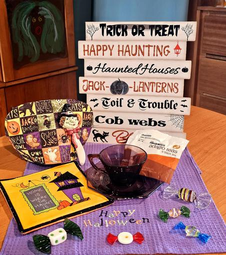A Halloween tea tableau in purple, orange, and green with Murano glass candies, a vintage purple glass cup & saucer, a purple tea towel embroidered with "Happy Halloween" in candy colors, a hot pad with a haunted house, cobwebs & bats and the phrase, "Home Is Where the Haunt Is". There's a patchwork tea cozy with various Halloween scenes, a ceramic butter spreader in the shape of a witch's head, and a tall wooden directional sign with boards that say, from the bottom up, Black Cats, Cob Webs, Toil & Trouble, Jack-O- Lanterns, Haunted Houses, Happy Haunting, Trick Or Treat, each with a little symbol such as a cat, a bat, a spider, etc. The tea is Chocolate black tea from Adagio. In the background is a glass cabinet door painted with a shrieking, yellow-eyed, red-mouthed ghoul face clutching at its green hair.