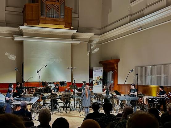 A view of a concert hall from the audience. A conductor faces the performers, a young Japanese percussion ensemble arrayed in a semicircle before her, all of them playing a variety of both typical and experimental percussion instruments.