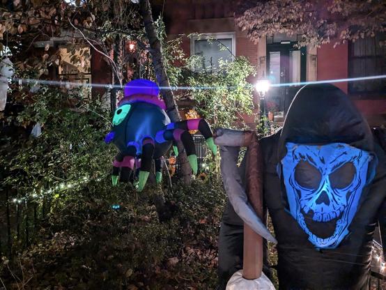 Photo of Halloween decorations in the evening in lawn in Carroll Gardens. An inflatable Grim Reaper is in the foreground, center right. We see in the background a large hanging inflatable spider from a tree. To the far left there is a white ghost from a sheed hanging in a tree. There is a lantern emitting a beam of light that looks like it is coming from the ghost.