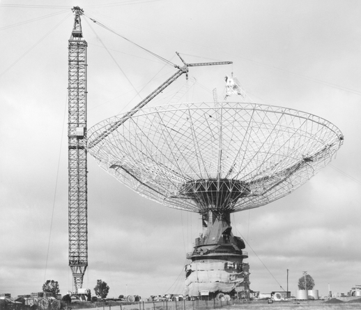 Old black and white photo of the Parkes radio telescope under construction with a crane nearby hoisting material onto the top of the skeletal dish structure