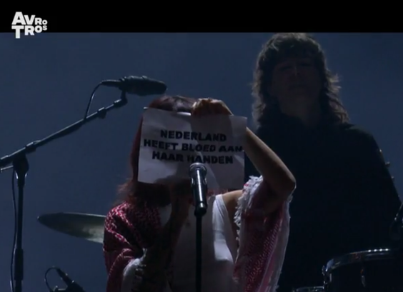 Uitsnede van een screenshot van de live-uitzending van het openingsconcert van 750 jaar Amsterdam met Sophie Straat die een stuk papier met de tekst "NEDERLAND HEEFT BLOED AAN HAAR HANDEN" in haar handen heeft.