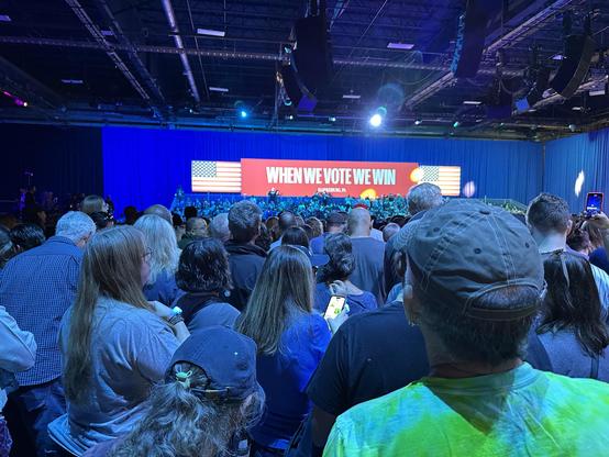 Color Harris rally photo - waiting for Kamala. Large When We Vote We Win sign over full bleacher seats. Crowded floor illuminated by blue and green glow.