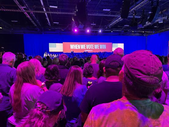 Color Harris rally photo - waiting for Kamala. Large When We Vote We Win sign over full bleacher seats. Crowded floor illuminated by red glow.