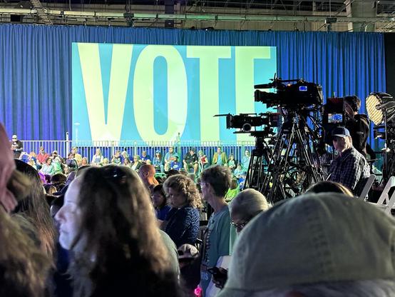 Color Harris rally photo - waiting for Kamala. Vote sign over full bleacher seats. TV cameras on a platform to the right, pointed over crowd, toward podium. illuminated by blue and green glow.