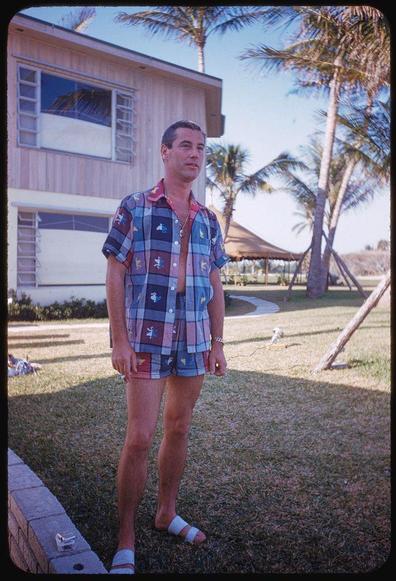 A color photograph of a man standing in front of a single-story wooden house with large windows and an air conditioning unit. The sky is clear, suggesting it's taken during the day under good weather conditions.

The subject appears to be wearing casual attire consisting of shorts adorned with colorful patches or badges on each leg, and a buttoned-up short-sleeve shirt that matches in design and color scheme as his shorts but has fewer buttons undone at the top. His right hand is partially visible, possibly gripping something small like sunglasses or jewelry.

He's standing on what looks to be an outdoor patio with grass surrounding it, indicating this photo was taken outside a residence during daylight hours.

In terms of background details, there are palm trees suggesting a warm climate and perhaps coastal location. The house seems dated, potentially from the early-to-mid 20th century based on architectural style. On closer inspection of his shorts, we can see some distinctive designs or emblems that may offer clues to his interests.

On one side lies an empty cigarette pack with 'Marlboro' branding visible on it and a small rectangular object beside him which could be either the Marlboro's lighter case or another item entirely. The setting suggests leisure time possibly in a residential backyard area near a beachfront given palm trees are often found along such locales.

The man is wearing white, casual sandals, imply [...]