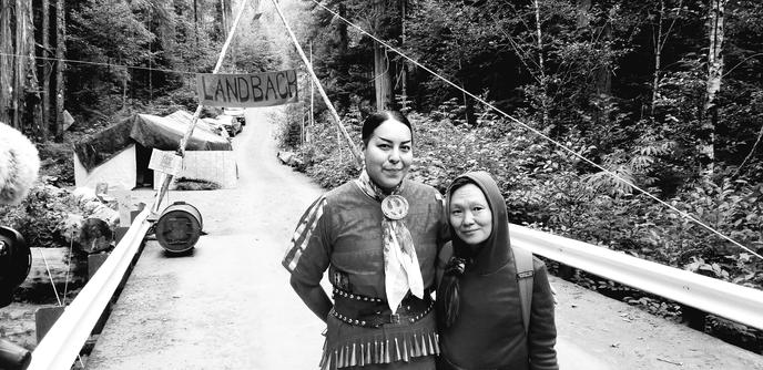 Me(in a hoodie with a backpack) & Silena(wearing her Ojibwe jingle dress), on bridge by landback resistance camp. We are surrounded by old growth forest.
Black and white photo.