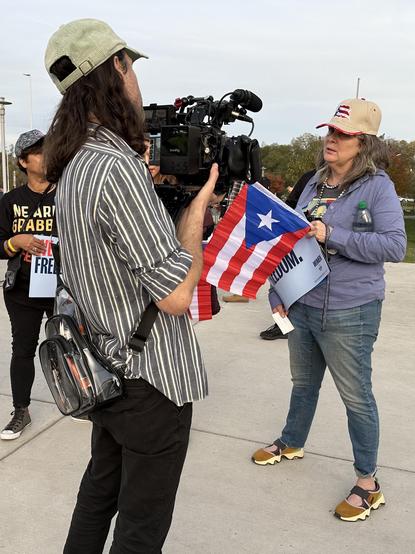 Color photo of reporter with a large video camera over his shoulder interviewing four Kamala Harris supporters who each carried a modestly sized Puerto Rican flag into the Rally. A photo shot from the reporter’s perspective.