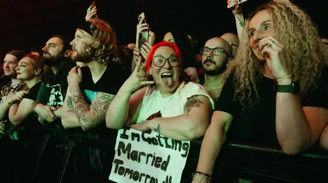 A group of people stand at the front row of a rock concert, waiting for the band. In the centre of the image is a person with red hair, sticking out their tongue and giving the good old rock "devil's horns" sign with their hand. They are smiling broadly and holding a large handwritten sign that says "I'm getting arried tomorrow!"