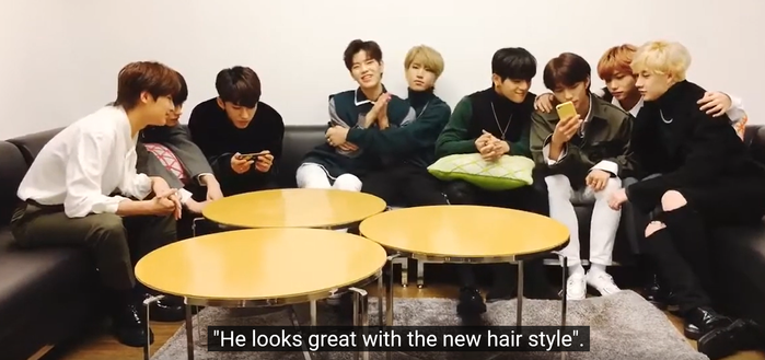 Stray Kids members sitting on a big u-shaped sectional couch during a livestream. Han is tightly hugging Seungmin, whose arms are trapped. Caption says: "He looks great with the new hairstyle."