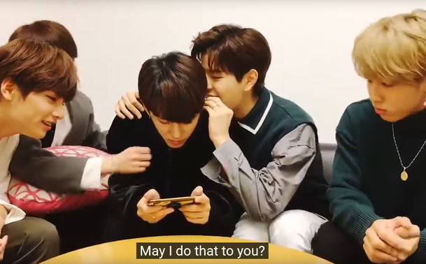 several stray kids members sit on a couch. Changbin looks at a phone he's holding in both hands. Seungmin has one arm around Changbin. He's holding Changbin's ear and leaning in to attempt to bite it. Caption says: May I do that to you?