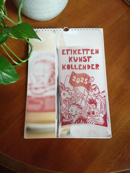 Ein Kalender auf einem Holztisch mit einer Grünpflanze im Hintergrund.

Auf dem Kalender ist eine Falsche von hinten abgebildet, sodass die Etikettenkunst zu erkennen ist. Darauf die 4 Maskottchen von Kollemate, die Zotrine-Ziege, der Kolle-Igel, der Kilimo-Hahn und das Kolla-Faultier, die zusammen 2 Fahnen halten. Auf der Forderten steht 2024. Über den Tieren die Schrift: "Etiketten Kunst Kalender".