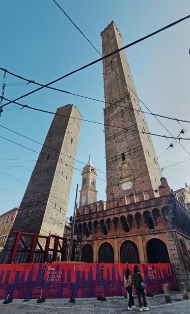 Saluti da Bologna.

This image shows the Two Towers of Bologna, known as the Asinelli Tower and the Garisenda Tower, located in Bologna, Italy. These medieval towers are a symbol of the city. The photo also includes a church in the background and features some construction barriers in the foreground.