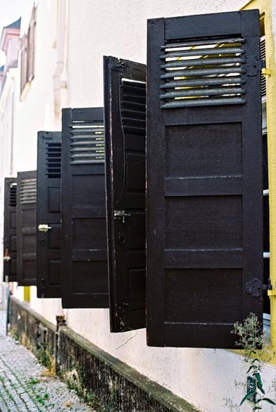 The picture shows a section of an old building. The façade is bright and plastered. Several windows are lined up next to each other. Each window is fitted with black wooden shutters. These shutters are divided into two parts and have a row of small, horizontal slats at the top. Some shutters are fully open and protrude at right angles to the wall. The shutters are fitted with simple black fittings. A narrow footpath with cobblestones can be seen in the foreground. A small plant grows against the wall.