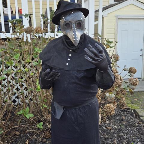 Photograph of a person in a long, black plague doctor costume shown facing frontward in front of a bush and a yellow house.