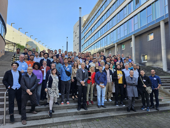Participants of the FAIR-DI Conference in Karlsruhe. There are roughly 80 Participants standing on the steps of a wide stair next to a huge building. Above, blue skies and nice weather.