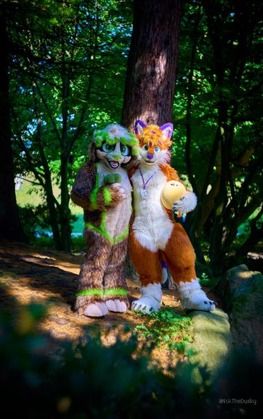 Fox und Gustl Flummox in Fullsuits stehen in einem Park vor einem Baustamm, im Schatten von dichtem Laub. Fox hält ein sehr rumdes Plüschhühnchen und Gustl einen Plüschbrokkoli.