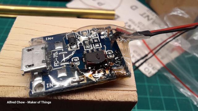 A postage stamp sized circuit board, it has a USB socket on the left, a chip and some surface mount components on the board, and red and black wires on the right.
The chip is overheated, melting through the heatshrink sleeving that was over the board, and some of the components have melted off, too.
There is burning and discolouration on the board.