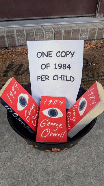 A black bowl decorated with orange Halloween pumpkins contains multiple copies of George Orwell's book 1984, each with a red cover featuring a stylized eye. Behind the books, a sign reads, 'ONE COPY OF 1984 PER CHILD.' The bowl is placed on a concrete ground near a brick step.