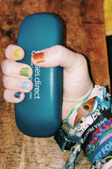 My hand holding a glasses case. The fingernails are all different colours; orange, yellow, green, light blue and slightly darker blue. There are multiple festival bands on my wrist.
