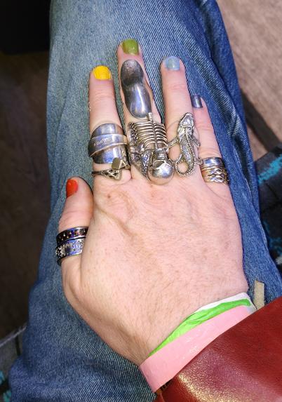 My hand resting on my lap. The fingernails are all different colours; orange, yellow, green, light blue and slightly darker blue. Each finger has multiple rings on it including a silver skeleton, a silver gecko and a flattened silver teaspoon ring.